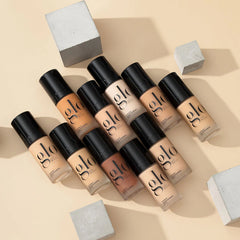 Verschiedene Glo Skin Beauty Liquid-Foundation-Flaschen in verschiedenen Farbtönen, diagonal mit Betonkuben arrangiert