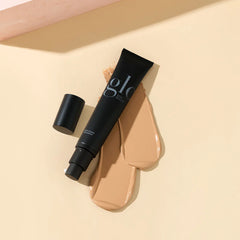 Schwarze Foundation-Tube, beschriftet mit Glo Skin Beauty und Skin Cream Foundation, mit zwei beigefarbenen Swatches auf hellem Hintergrund