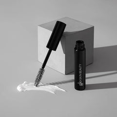 Schwarze Mascara-Tube mit Glo Skin Beauty Logo, Mascara-Bürste auf weißem Cream-Smear platziert, daneben ein grauer geometrischer Block – Produktdarstellung der Glo Skin Beauty Mascara.