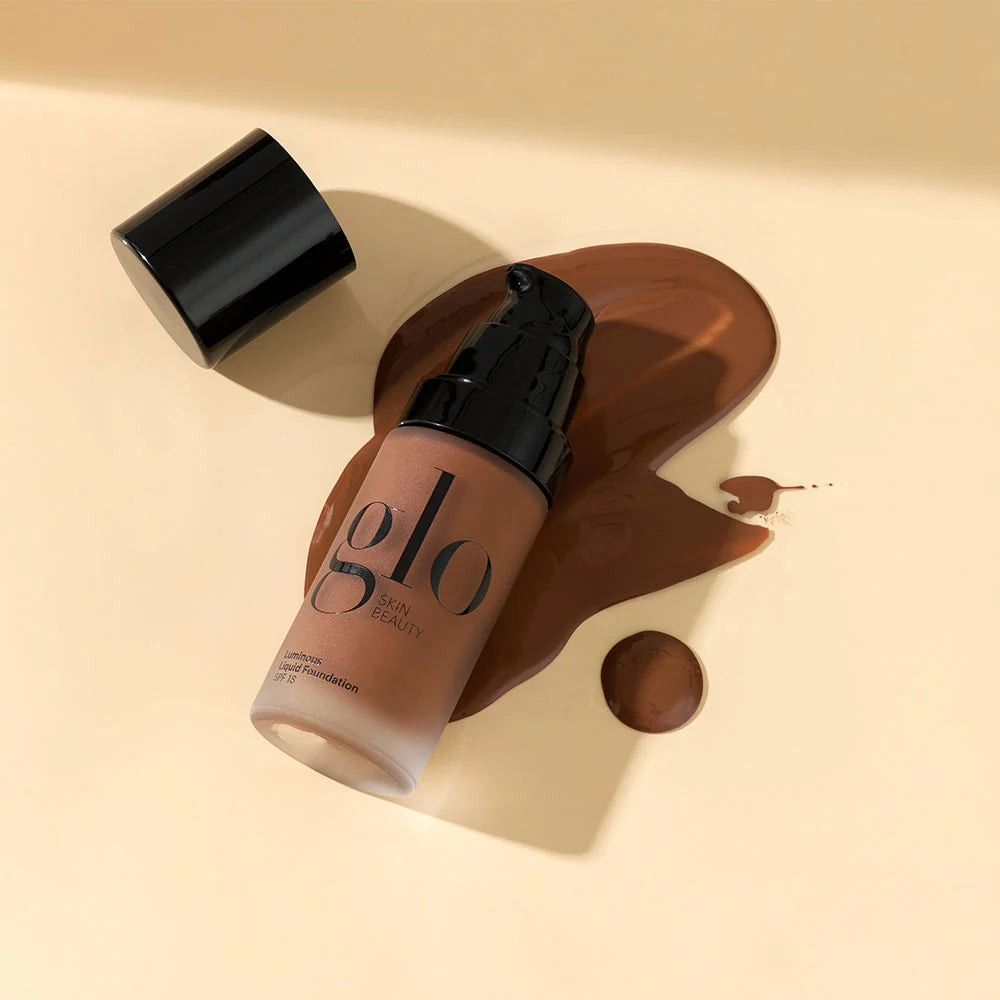 Glo Skin Beauty Luminous Liquid Foundation SPF 18 Flasche mit verschüttetem mittel-dunklem Farbton auf beigefarbenem Hintergrund