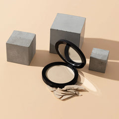 Geöffnetes schwarzes Kompakt mit beigefarbenem gepresstem Gesichtspuder und verstreuten Puderflocken, umgeben von drei Betonkuben auf beigefarbenem Hintergrund