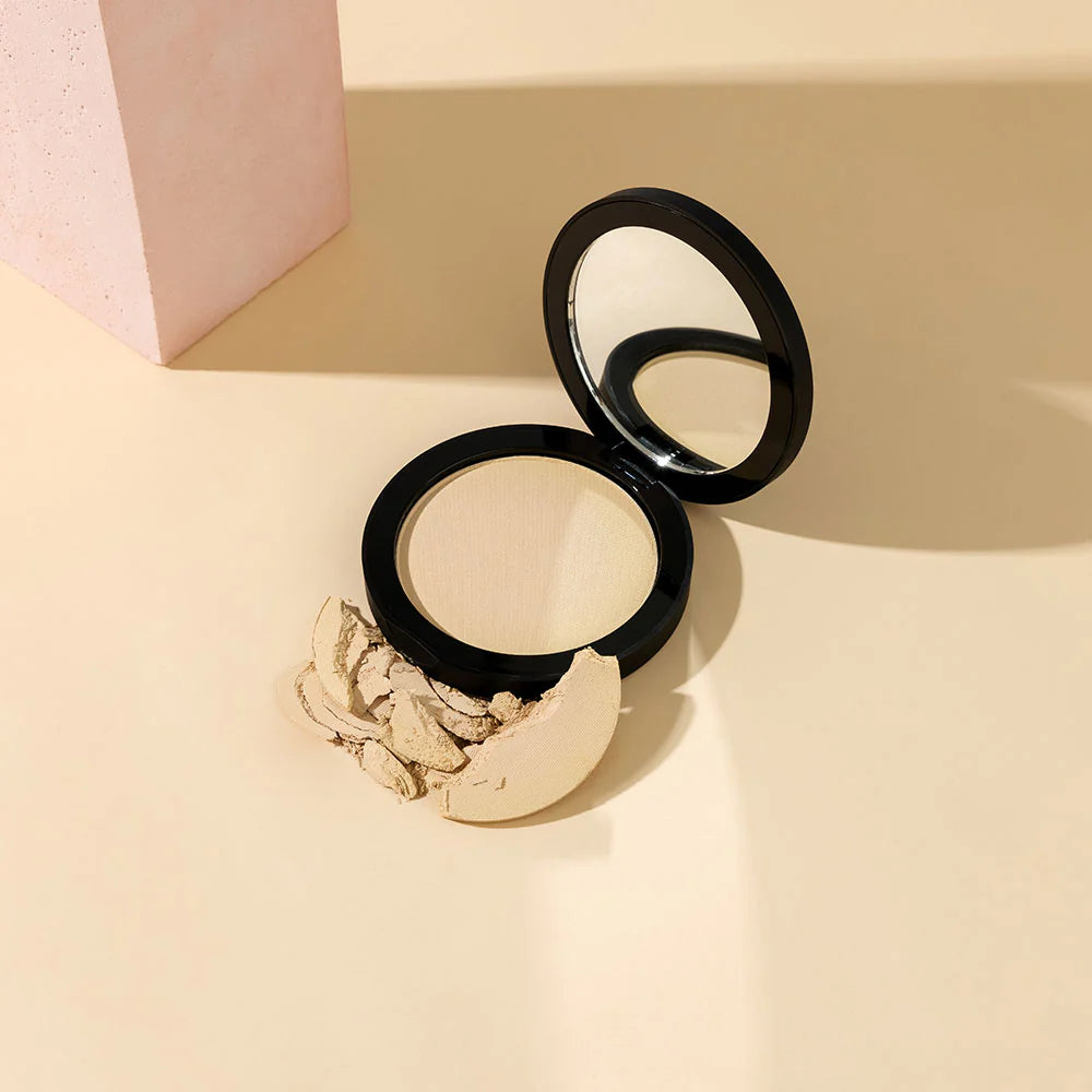 Geöffneter Kompaktpuder mit Spiegel und zerstoßenem hellbeigen Puder auf beigefarbener Oberfläche