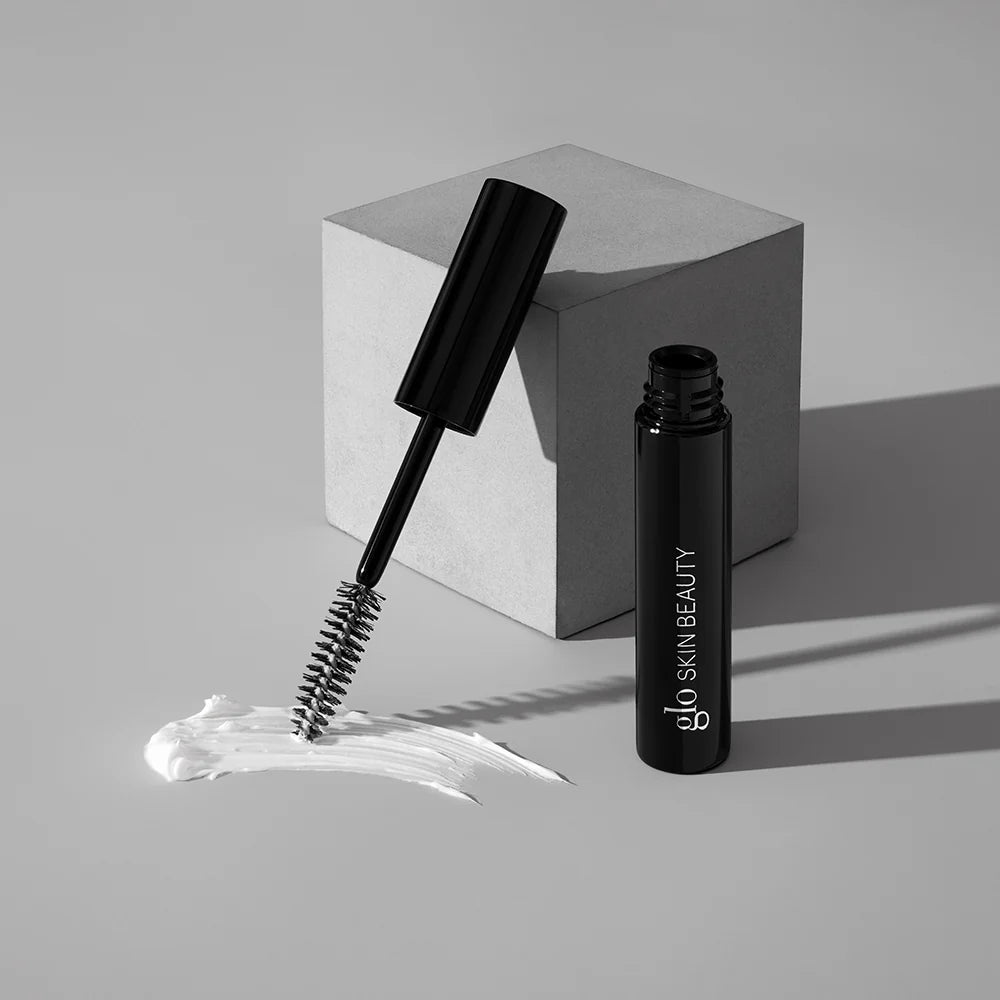 Schwarze Mascara-Tube mit Glo Skin Beauty Logo, Mascara-Bürste auf weißem Cream-Smear platziert, daneben ein grauer geometrischer Block – Produktdarstellung der Glo Skin Beauty Mascara.